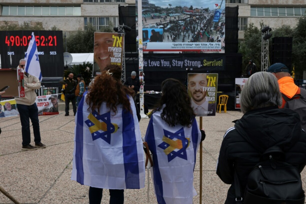 Des femmes drapées d'un drapeau israélien dont l'une tient une pancarte avec la photo d'Or Levy, l'un des otages devant être libérés samedi à Gaza, regardent un écran géant installé sur une place de Tel-Aviv et transmettant des images des préparatifs de libérations