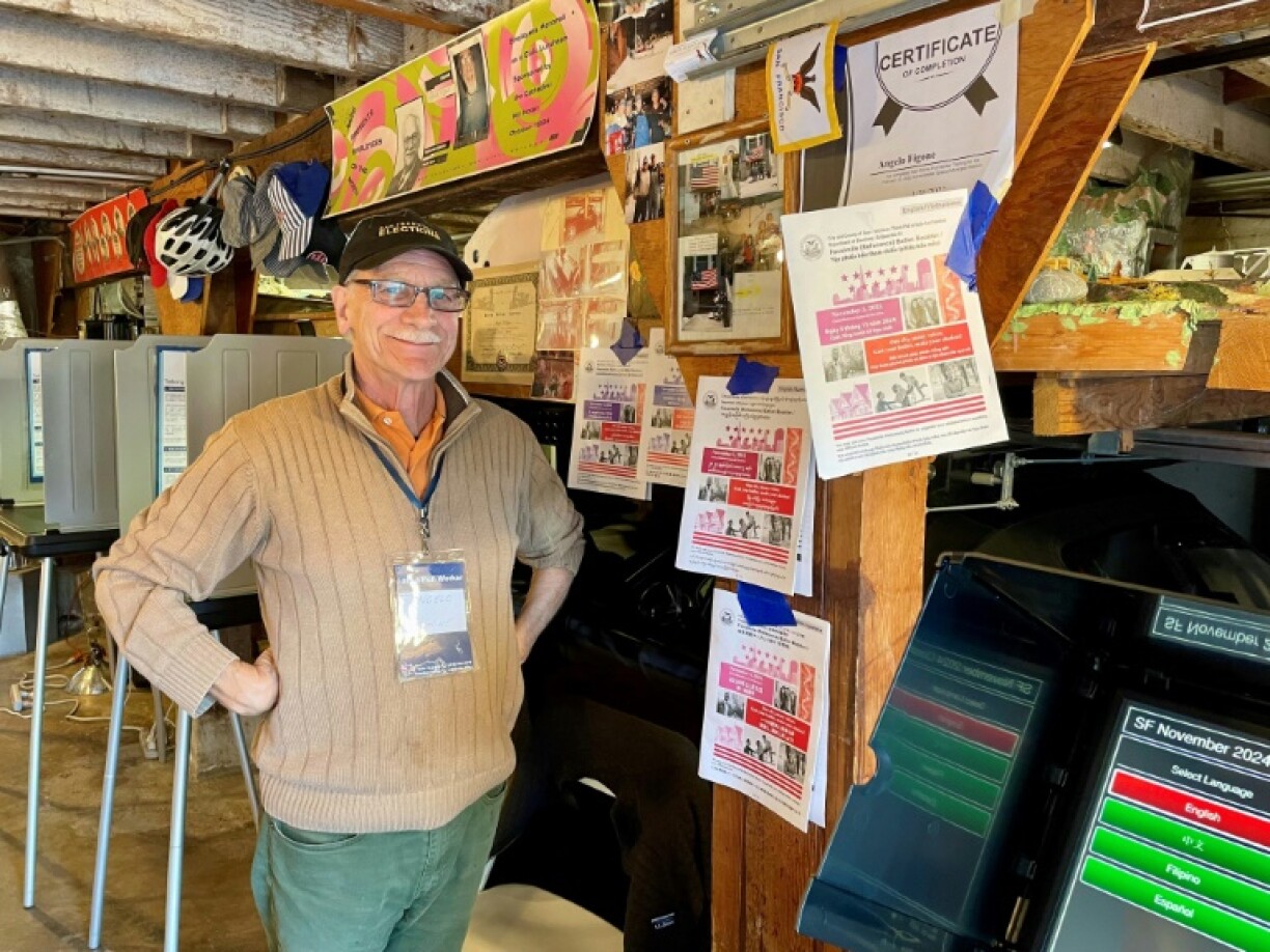 Angelo Figone, un retraité de 76 ans, pose le 5 novembre 2024 à San Francisco dans son garage transformé en bureau de vote pour la journée, devant les isoloirs, les machines et notices officielles, et des photos de précédentes élections