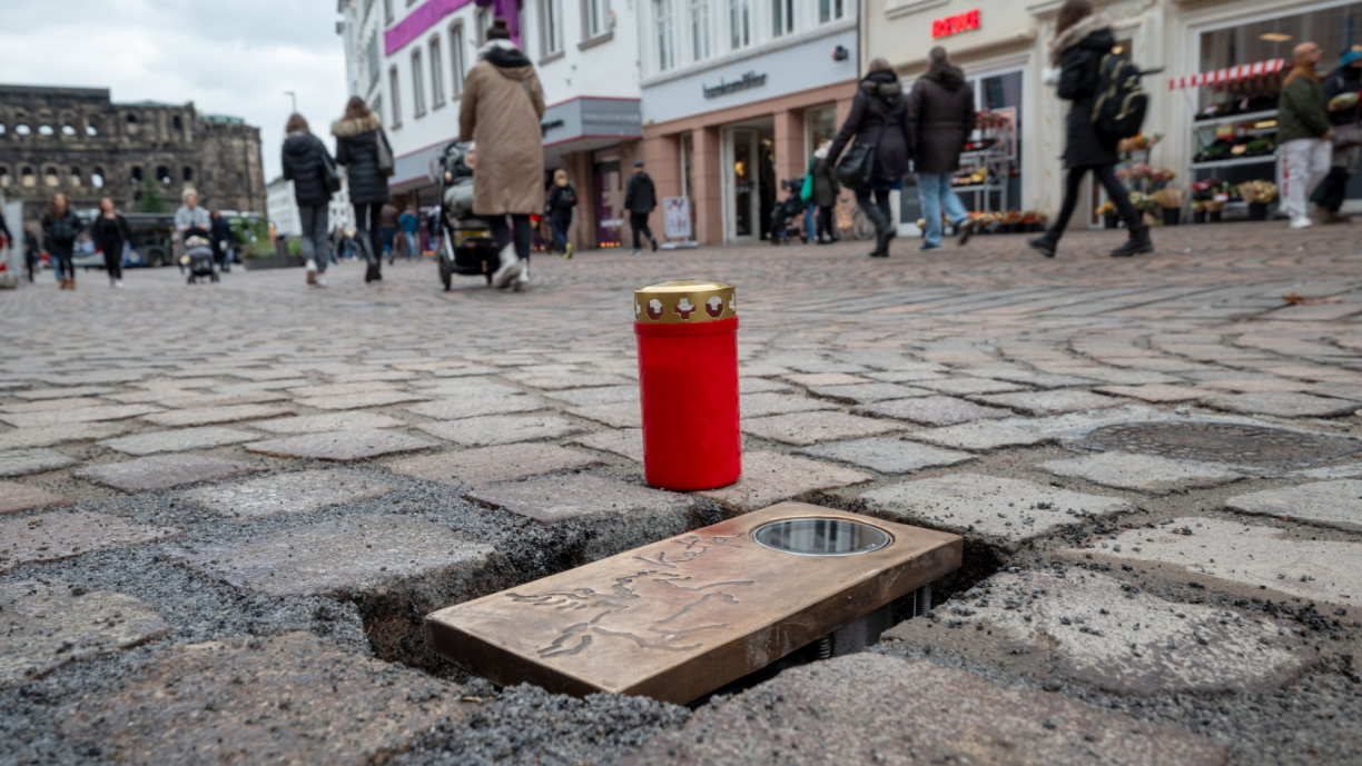 17 November 2023, Rhineland-Palatinate, Trier: A memorial plaque for one of the victims of the rampage on December 1, 2020 is embedded in the pavement near the Porta Nigra (back l) for presentation. The memorial plaques were designed by Trier artist Guy Charlier in close consultation with the bereaved and will be permanently installed in the next few days at the five locations in the pedestrian zone where people were killed by the driver of the amok. The plaques and lighting will be laid in prepared places in the pavement.