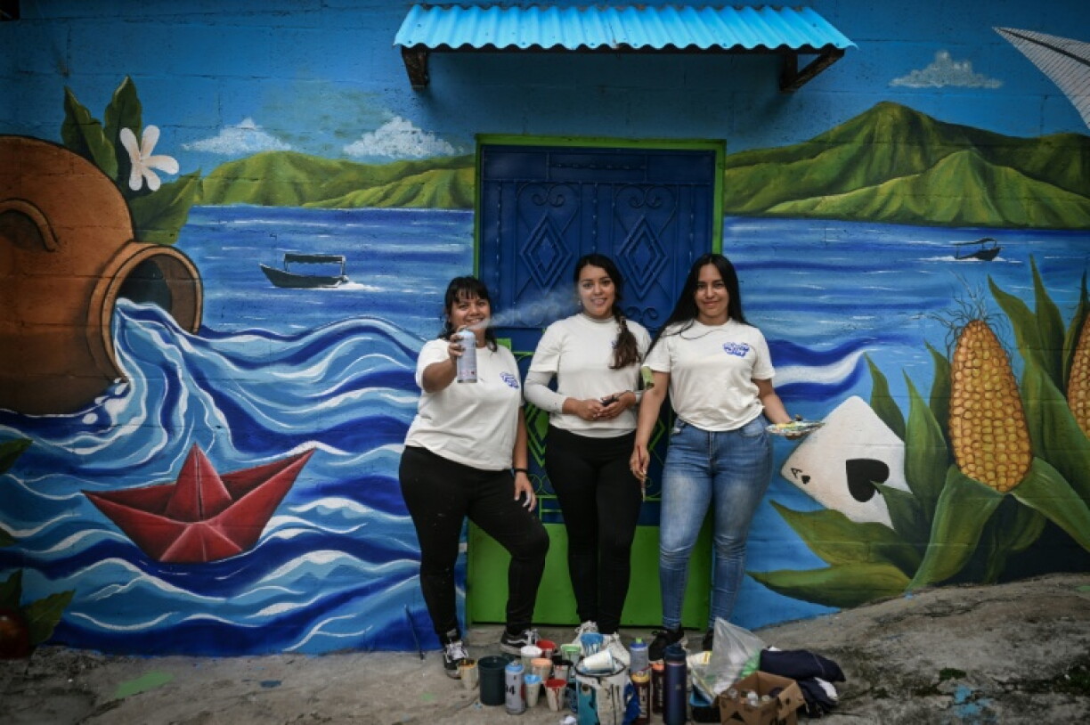 Las artistas salvadoreñas Patricia Vega (izquierda), Elizabeth Guardado (centro) y Marcela Alvarenga (derecha) posan frente a una casa cuya fachada están decorando con el mural «Identidad», en la comunidad 10 de Octubre de San Marcos, El Salvador, el 2 de octubre de 2026