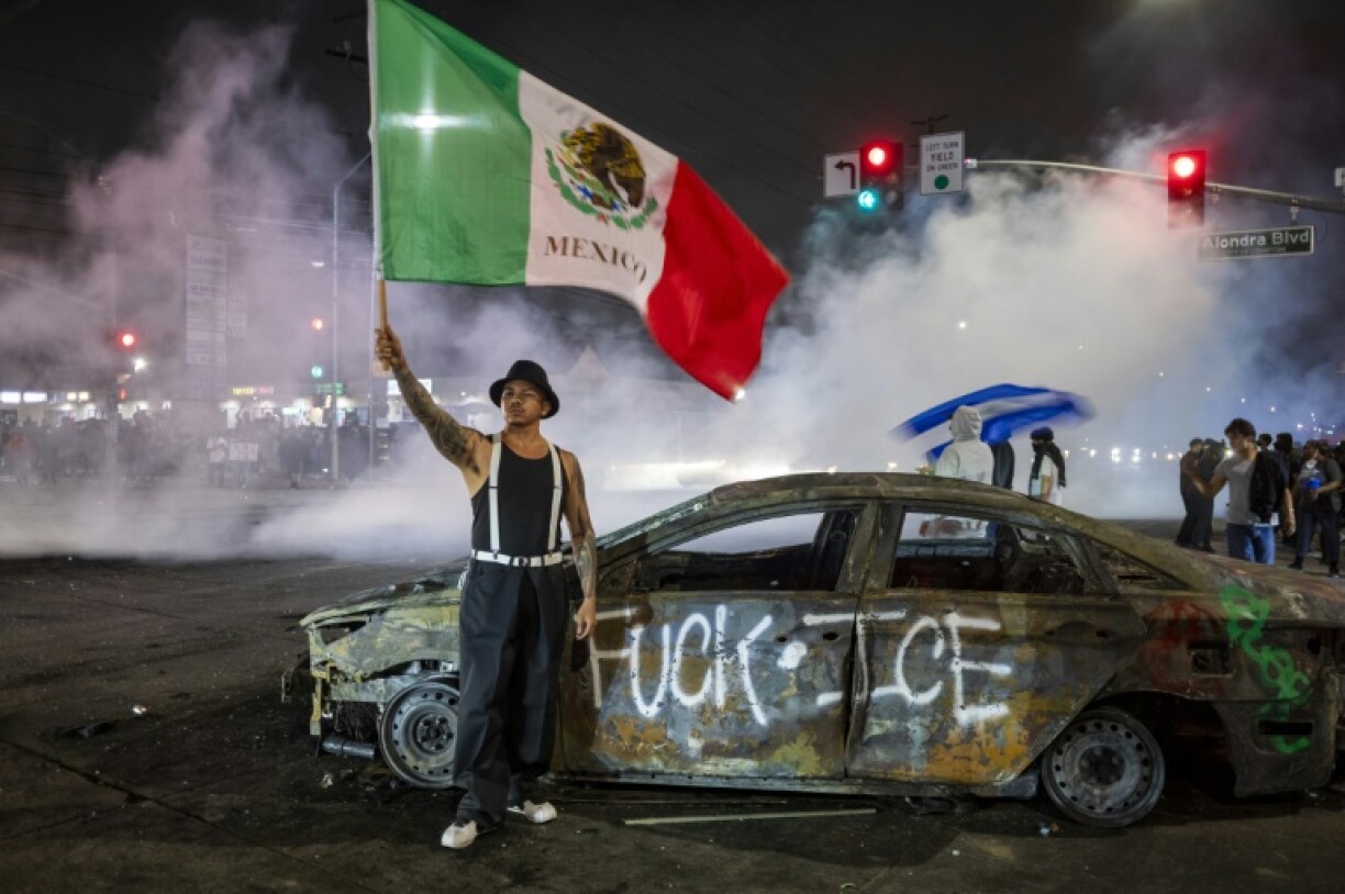 A protester holding a Mexican flag stands in front of a burnt-out car that had been sprayed with a slogan against the immigration enforcement agency