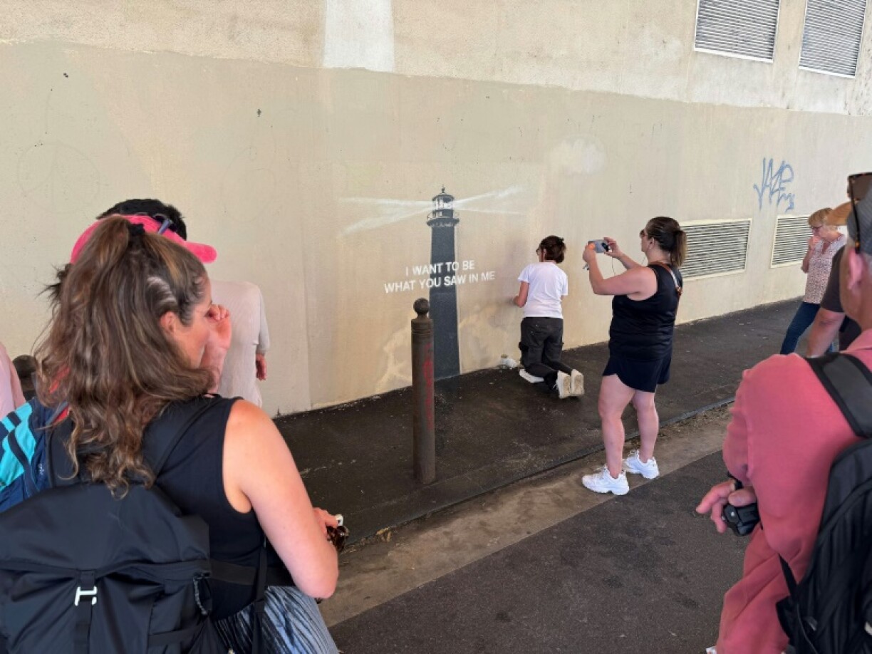 Des passants prennent des photos pendant que la peintre en décor du patrimoine, Agnès Perrone, répare une œuvre d'art de l'artiste de rue Banksy représentant un phare sur la façade d'un bâtiment, après qu'elle a été vandalisée pendant la nuit à Marseille, le 1er juin 2025