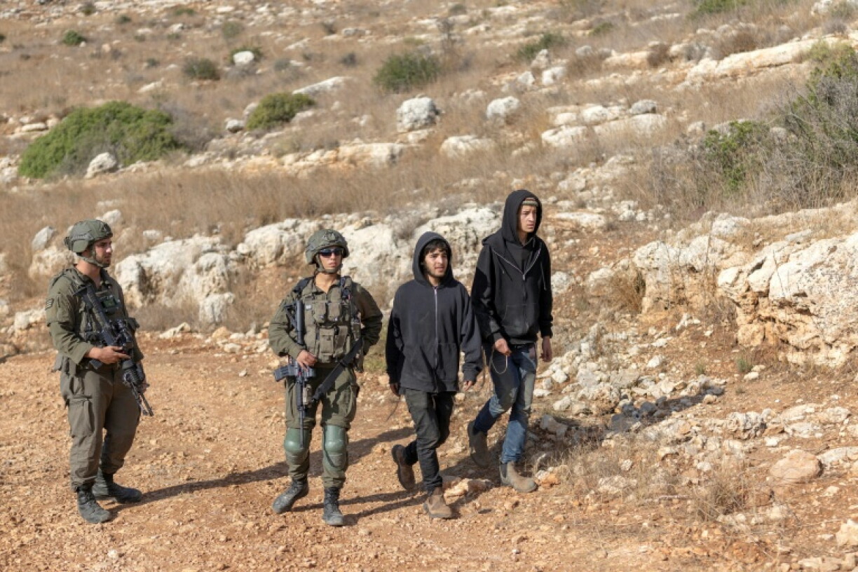 Israeli soldiers look on as settlers walk along a dirt road where Palestinian farmers are harvesting their olive trees in the West Bank village of Deir Abu Mashal