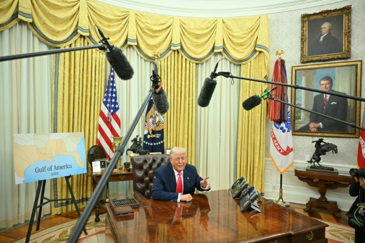 US President Donald Trump at the Resolute Desk, flanked by a map showing the