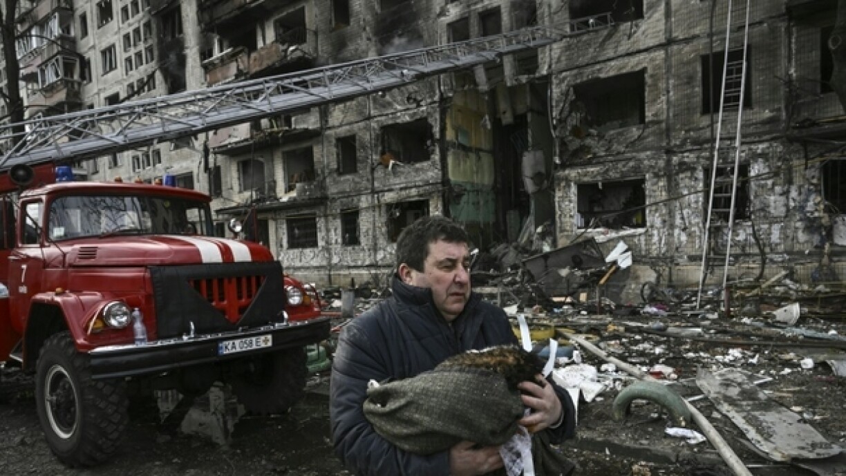 E Mann mat senger doudeger Kaz am Aarm virun engem zerstéierte Wunnquartier.