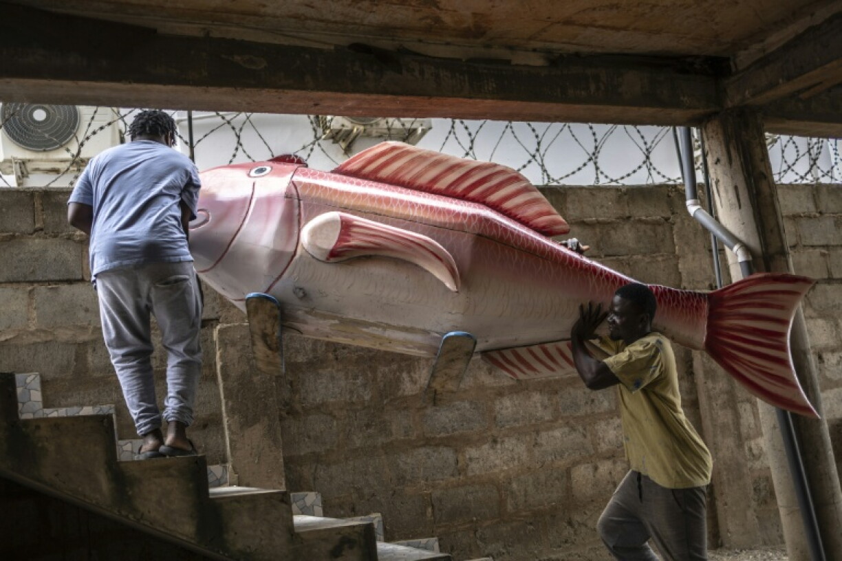 Derrick Anang (à gauche) et son frère Lawrence Anang, tous deux fabricants de cercueils, transportent un cercueil en forme de poisson, conçu en référence au métier de pêcheur du défunt, dans leur atelier de menuiserie à Accra, au Ghana, le 17 août 2026