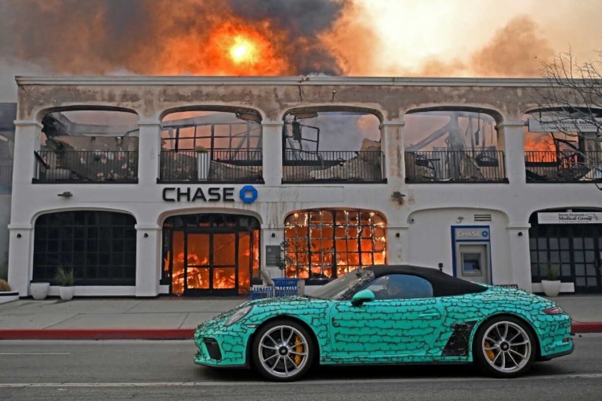 A sports car decorated with Christmas lights drives past a burning bank during the Palisades Fire