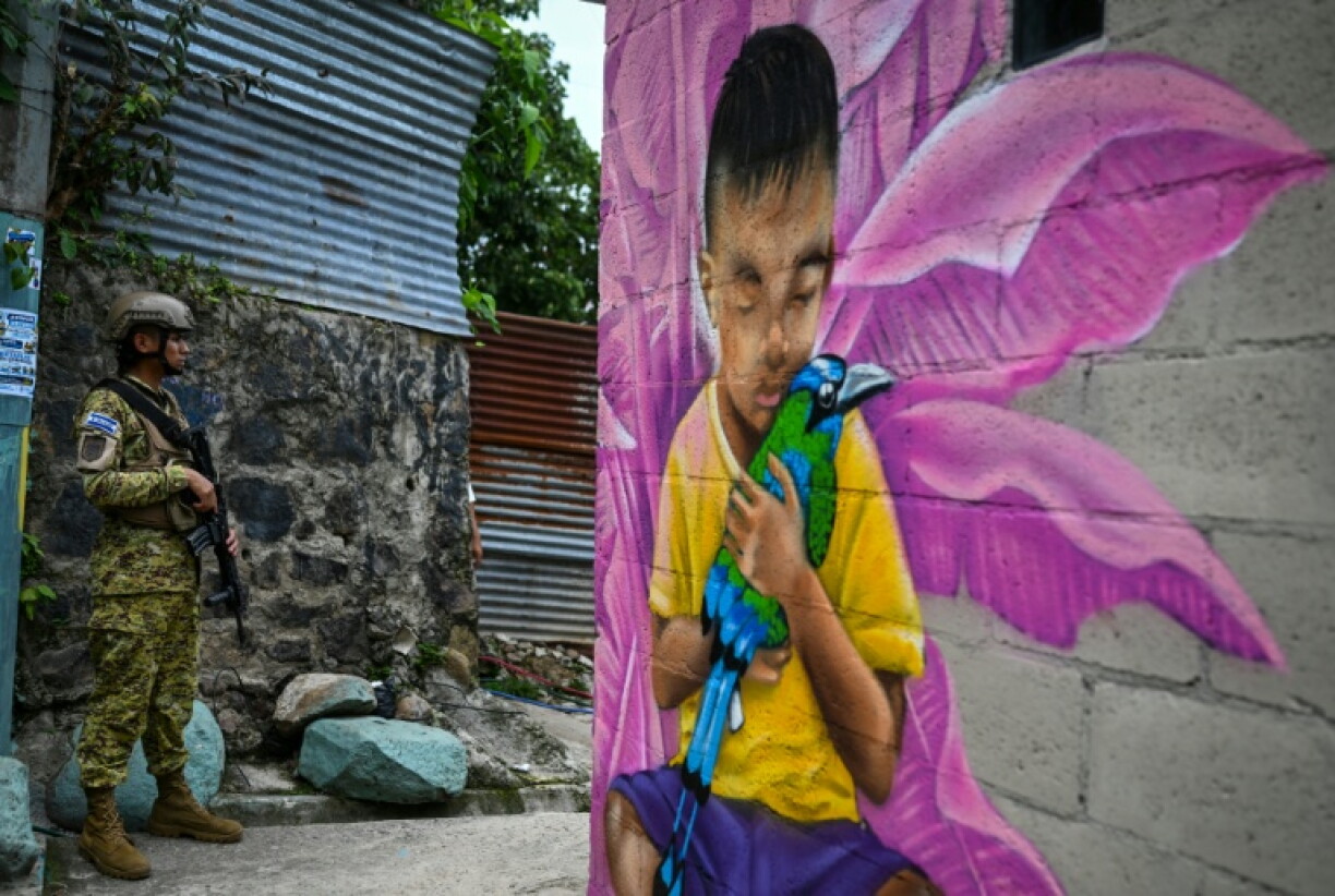 A Salvadoran soldier stands guard in a narrow passage in the 10 de Octubre community, in front of a mural painted by artist Carlos Clímaco, in San Marcos, El Salvador, on October 2, 2026. The artistic project aims to transform the identity of a community historically affected by gang violence.