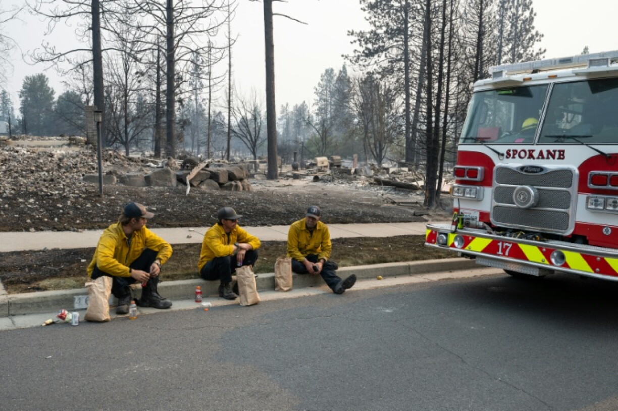Trois membres du service incendie de Spokane assis sur le bord du trottoir devant des maisons d'habitation incendiées, dans le quartier de Balboa à Spokane (Washington), le 2 août 2026